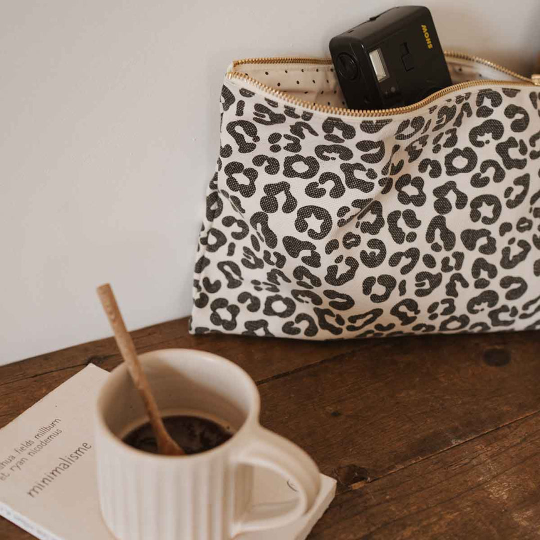 Lili Flat Pouch in leopard greige print, perfect for makeup and toiletries, on a wooden table near a coffee cup.