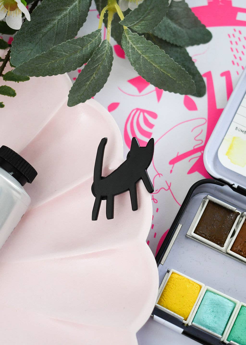Black cat-shaped enamel pin on a pink plate beside watercolor paints and flowers