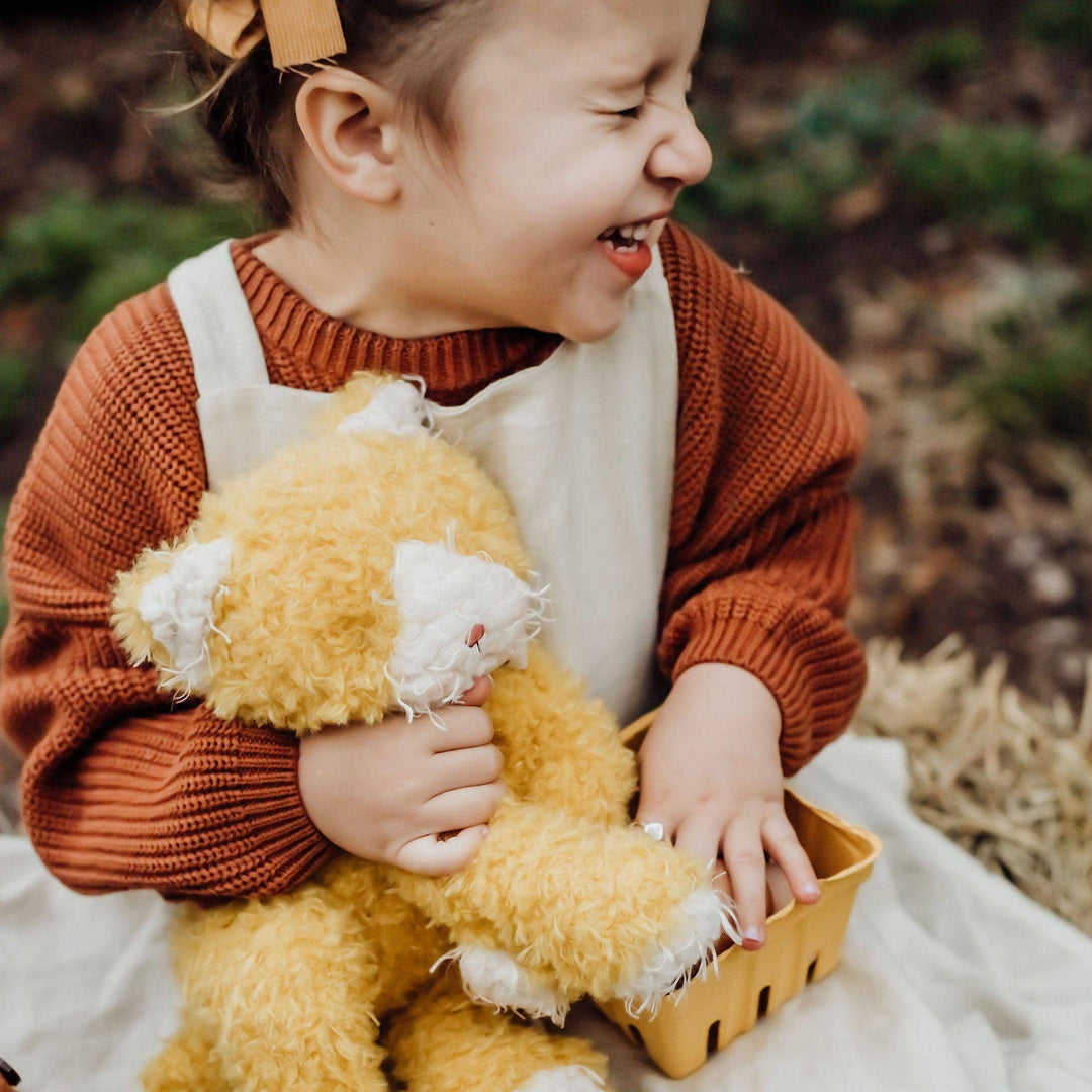 Child holding yellow Alley Cat stuffed animal with scruffy fur, cream muzzle, and polka dot scarf in an outdoor setting.