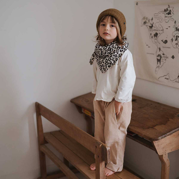 Child in a cozy outfit with a leopard print scarf and beanie, standing on a wooden bench in a bright room.