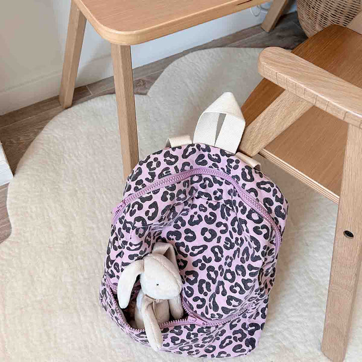 Kids leopard print backpack in pink with plush rabbit toy beside wooden chair, showcasing playful and stylish design.