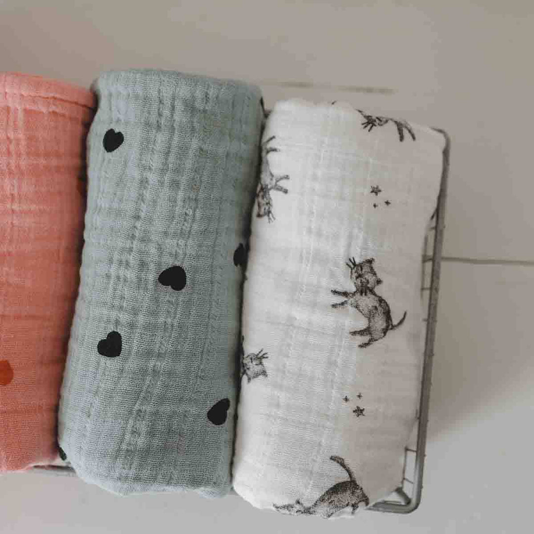 Three cotton muslin swaddles in coral, teal with hearts, and white with a cat print, displayed in a basket.