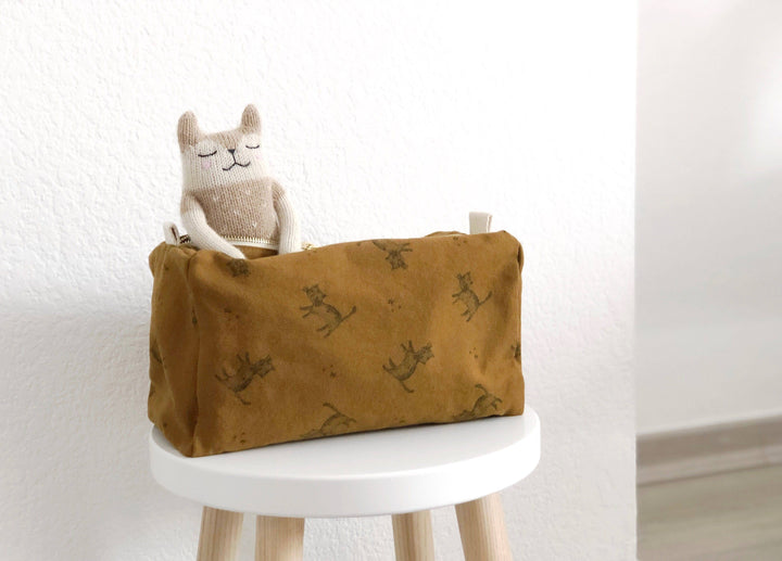 Kids' toiletry bag with cat print in cumin color, placed on a white stool alongside a decorative plush toy.