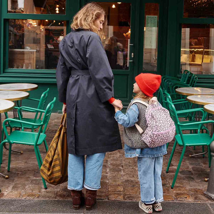 A woman in a black coat and a child in a red beanie hold hands while standing outdoors near tables.