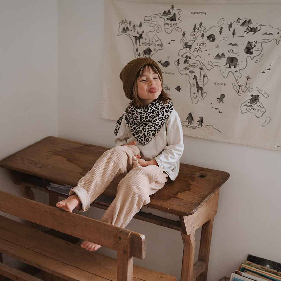 Child sitting on a wooden table wearing a leopard print scarf and beanie, with a world map tapestry in the background.