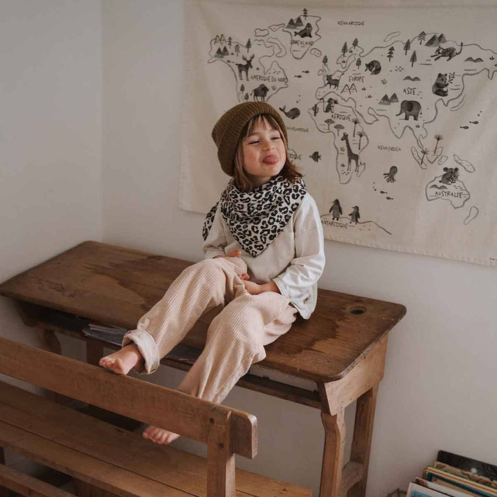 Child sitting on a wooden table wearing a leopard print scarf and beanie, with a world map tapestry in the background.