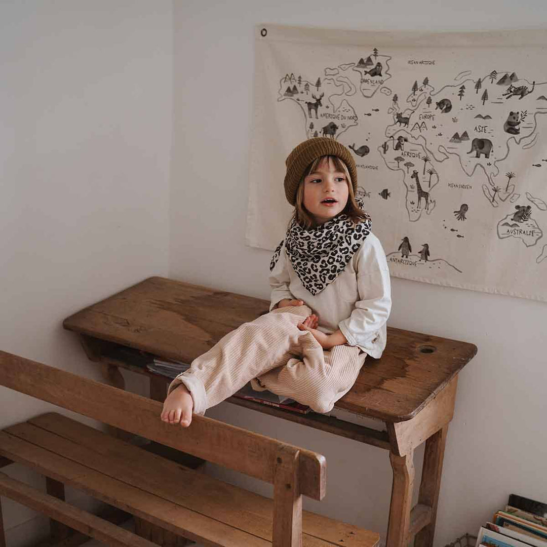 Child wearing a leopard print scarf and beanie, sitting on a wooden bench in a cozy room with a map decor.