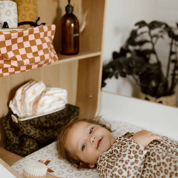 Vic Toiletry Bag in olive leopard print on a shelf. Child lying on changing table, surrounded by organized baby essentials.