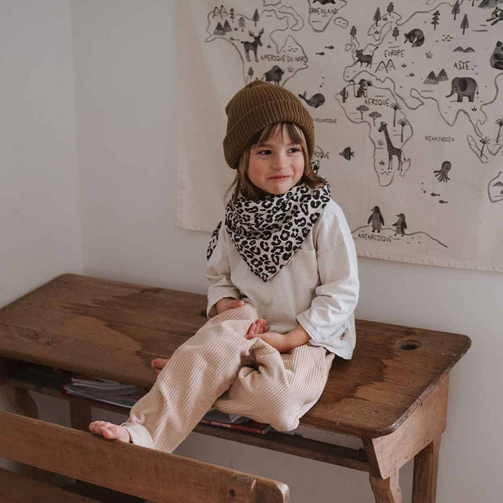 Child sitting on a wooden desk wearing a brown beanie and a leopard print scarf, with a playful expression.