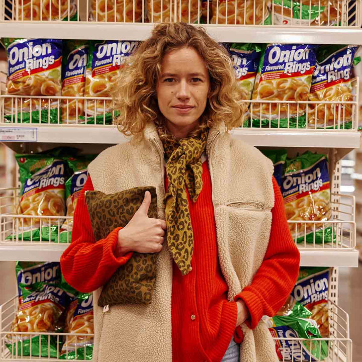 Woman holding Lili Flat Pouch in olive leopard print in front of a snack aisle with onion rings.