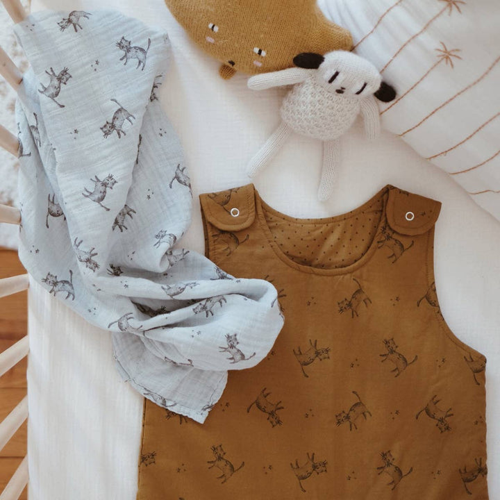 Bianca blue cat swaddle blanket with baby outfit and toys in a crib setting, showcasing 100% cotton muslin fabric.