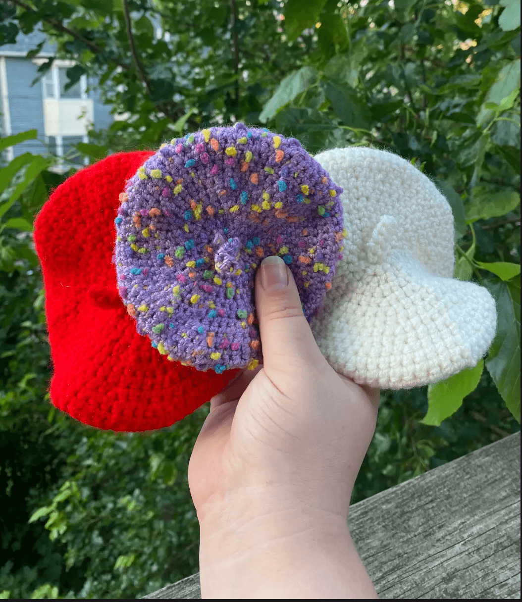 Hand holding three colorful crocheted cat beret hats in red, purple, and white with a blurred green background.