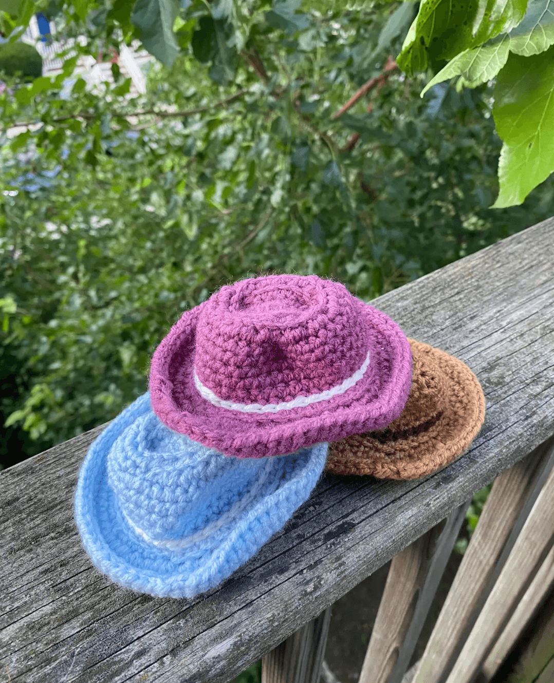 Handcrafted crocheted cat cowboy hats in purple, blue, and brown displayed on a wooden railing amidst greenery.