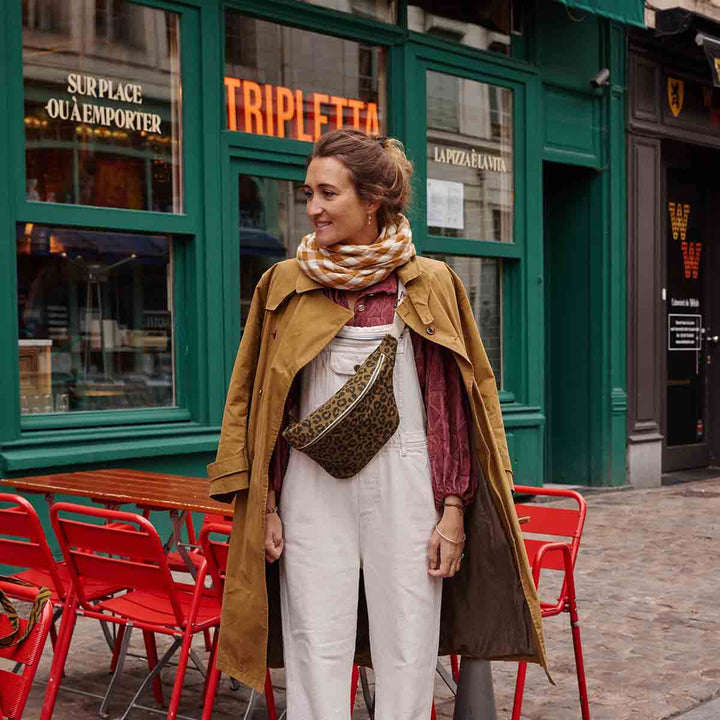 Woman in stylish outfit with leopard olive bum bag, standing outside a café with green walls and red chairs.