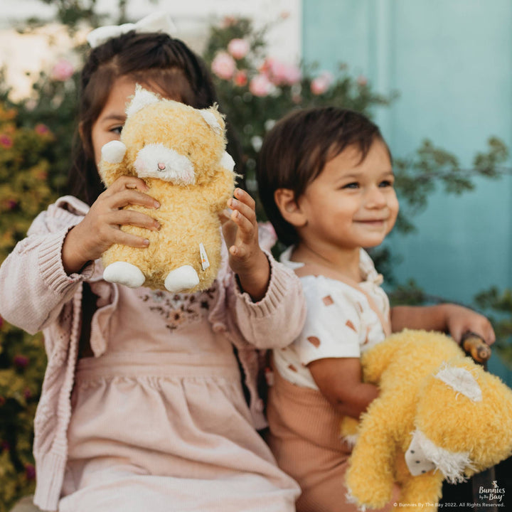 Two children holding and cuddling mustard yellow Wee Alley Cat stuffed animals outdoors.