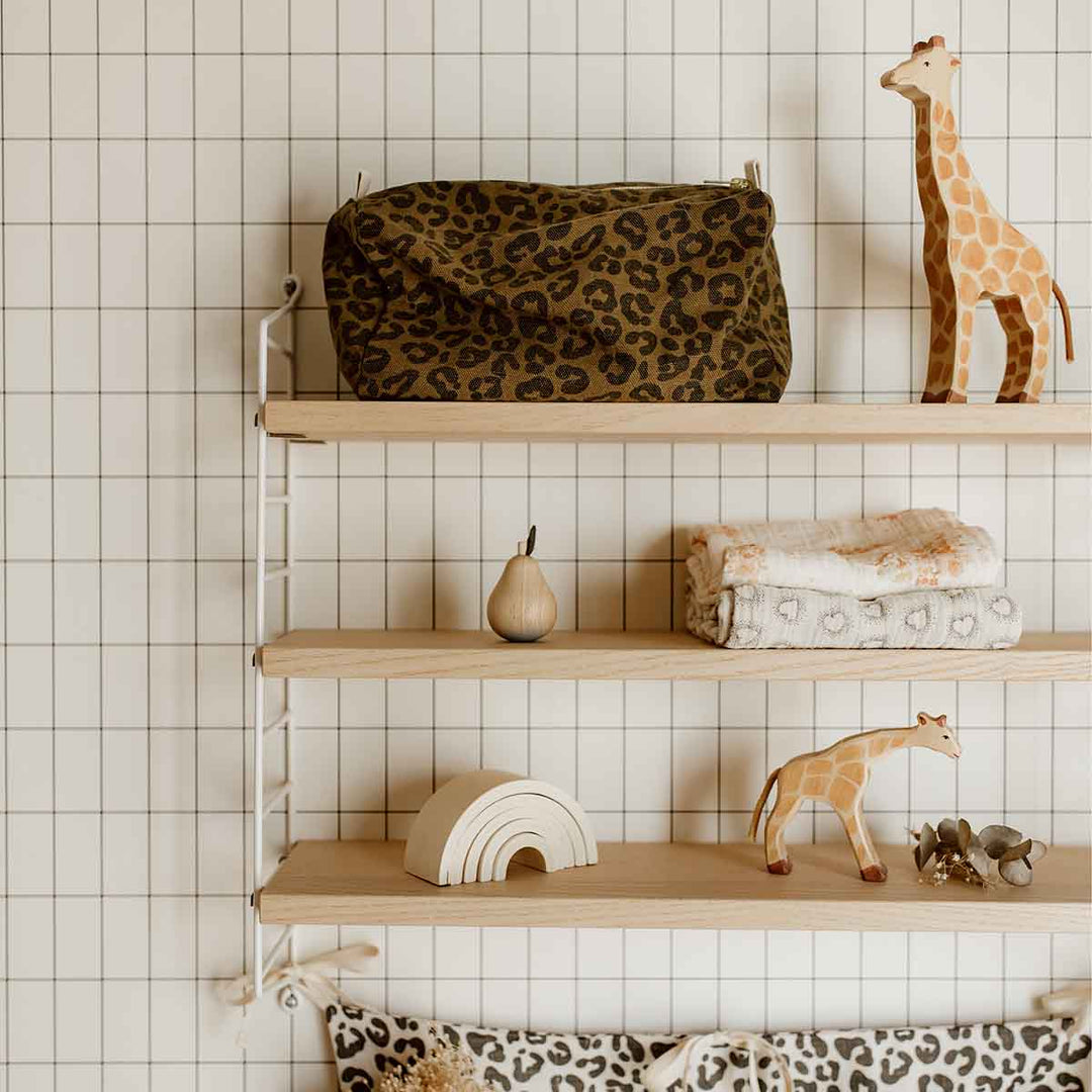 Vic Toiletry Bag in olive leopard print placed on a wooden shelf with children's toys and blankets.