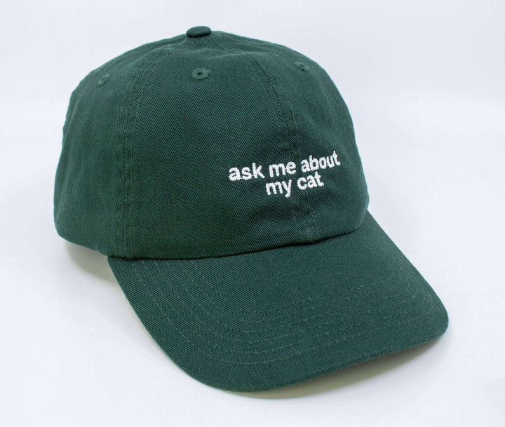Green embroidered hat with 'ask me about my cat' text, adjustable and made from 100% cotton.