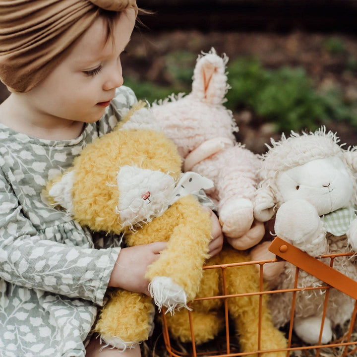 Child holding Alley Cat stuffed animal with mustard yellow fur and scraggly cream muzzle alongside other plush toys in a basket.